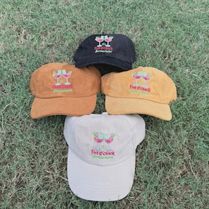 May include: Five corduroy baseball caps in black, tan, and off-white. Each cap features an embroidered cocktail design with the text "Five O'Crack Somewhere". The caps are arranged on a bed of green grass.
