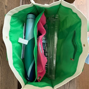 May include: A large open tote bag with a cream-colored exterior and a bright green interior lining. The bag contains a blue cylindrical tube, a pink zippered pouch with text, and several clear plastic rectangular frames organized inside.