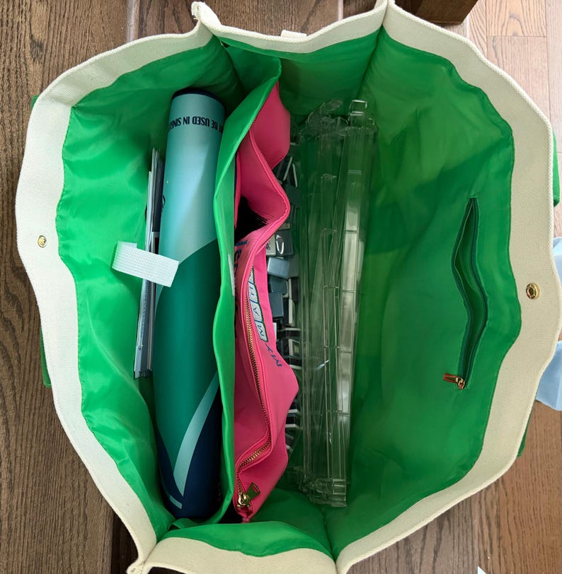 May include: A large open tote bag with a cream-colored exterior and a bright green interior lining. The bag contains a blue cylindrical tube, a pink zippered pouch with text, and several clear plastic rectangular frames organized inside.