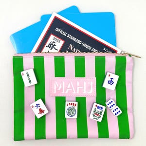 May include: A pink and green striped Mahjong pouch with a zipper, containing a booklet and blue cards. Several Mahjong tiles and dice are scattered around the pouch. The pouch has the word "MAHJ" on it.