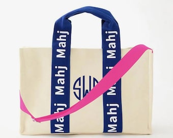 May include: A cream-colored canvas tote bag featuring two dark blue vertical straps with the repeating white text "Mahj" printed on them. A dark blue circular monogram logo is centered between the straps, and a bright pink crossbody strap is attached.