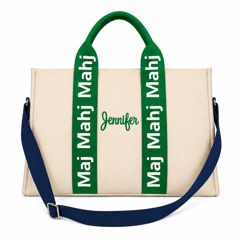 May include: A cream-colored tote bag featuring two green vertical straps with the white text "Maj Mahj Mahj" printed on them. The center of the bag displays the name "Jennifer" in green script. A dark blue adjustable shoulder strap is attached.