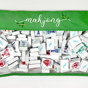 May include: A clear, zippered pouch filled with white mahjong tiles. The green top of the pouch has the word "mahjong" in white script, with two green crocodile illustrations. The tiles feature various colorful illustrations and blue mustache symbols.