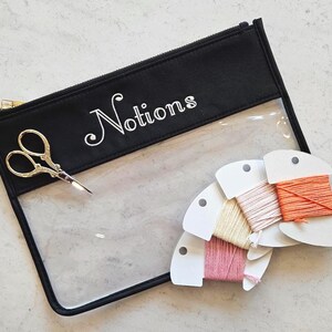 May include: A black zippered pouch with a clear front panel and the word Notions printed in white. The pouch contains a pair of gold-toned embroidery scissors and four white cardboard bobbins wrapped in pink, yellow, peach, and orange thread.