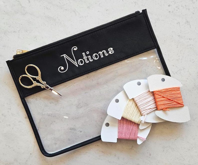 May include: A black zippered pouch with a clear front panel and the word Notions printed in white. The pouch contains a pair of gold-toned embroidery scissors and four white cardboard bobbins wrapped in pink, yellow, peach, and orange thread.