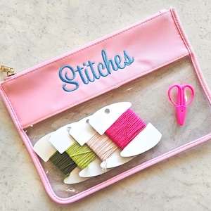 May include: A rectangular clear vinyl pouch with a pink border and a gold zipper. The word Stitches is embroidered in blue thread on the pink top panel. Inside are three white cardboard bobbins wrapped in green, beige, and pink thread, plus pink scissors.
