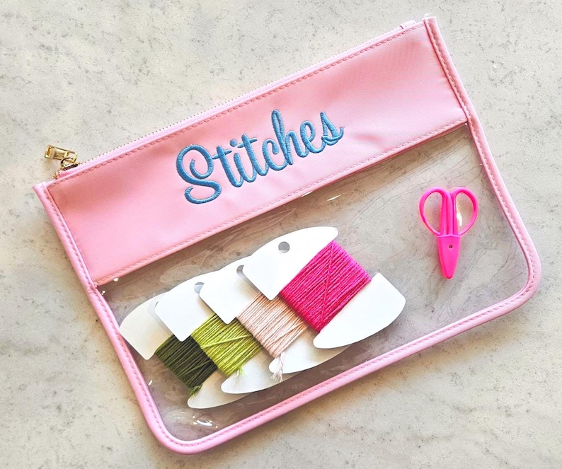 May include: A rectangular clear vinyl pouch with a pink border and a gold zipper. The word Stitches is embroidered in blue thread on the pink top panel. Inside are three white cardboard bobbins wrapped in green, beige, and pink thread, plus pink scissors.