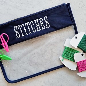 May include: A navy blue zippered pouch with the word STITCHES embroidered in white. The pouch features a clear plastic front panel. Included are a pair of pink thread snips, a pair of green thread snips, and three spools of embroidery floss.