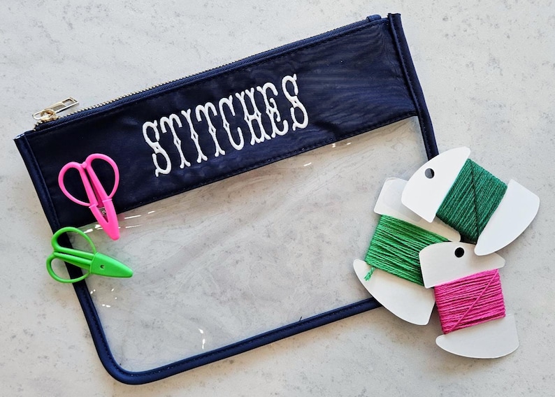 May include: A navy blue zippered pouch with the word STITCHES embroidered in white. The pouch features a clear plastic front panel. Included are a pair of pink thread snips, a pair of green thread snips, and three spools of embroidery floss.