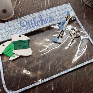 May include: A clear vinyl project bag with a blue and white floral fabric trim featuring the word Stitches embroidered in blue script. Inside are two green thread bobbins, two skeins of embroidery floss in blue and white, and small scissors.