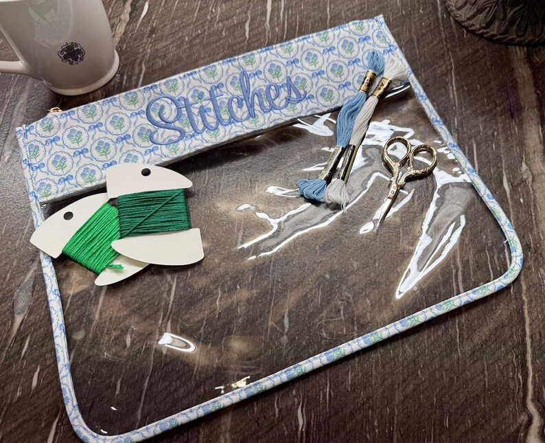 May include: A clear vinyl project bag with a blue and white floral fabric trim featuring the word Stitches embroidered in blue script. Inside are two green thread bobbins, two skeins of embroidery floss in blue and white, and small scissors.