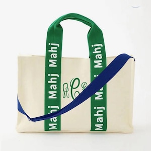 May include: Cream-colored tote bag with green handles and a blue shoulder strap. The green handles have the text "Mahj Mahj" printed in white. The bag features a green monogram and is designed for carrying items.
