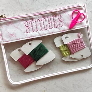 May include: A clear, zippered pouch with the word "STITCHES" embroidered in pink. Inside are spools of green, pink, and yellow thread, along with a pair of pink scissors. The pouch has a pink and white bow pattern.