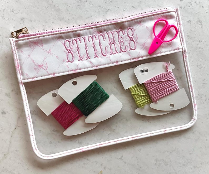 May include: A clear vinyl zipper pouch with a white fabric top featuring a pink bow pattern and the embroidered word STITCHES. Inside are four white cardboard bobbins wrapped in green, pink, and yellow embroidery floss, plus pink scissors.