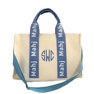 May include: A cream-colored tote bag featuring two light blue vertical straps with the repeated white text Mahj. The center of the bag displays a circular monogram with the letters SWC in light blue. It includes a matching light blue shoulder strap.