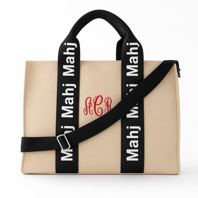 May include: A beige rectangular tote bag featuring black handles and a black shoulder strap. The black straps are printed with the white text Mahj Mahj Mahj. A red embroidered monogram with the letters ACB is centered on the front of the bag.