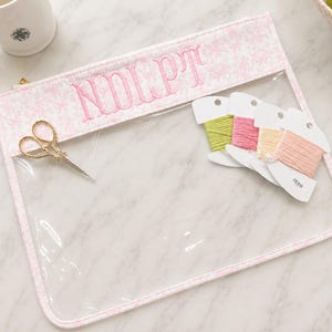 May include: A clear vinyl zipper pouch with a pink floral fabric border and the embroidered letters "NDLPT" in pink thread. Inside the pouch are gold-toned embroidery scissors and four spools of embroidery floss in green, pink, yellow, and peach.