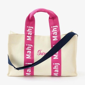 May include: A cream-colored canvas tote bag featuring two bright pink vertical straps with the white text "Mahj" printed repeatedly. A dark navy blue adjustable shoulder strap is attached, and the script name "Tara" is embroidered in pink.