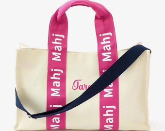 May include: A cream-colored canvas tote bag featuring two bright pink vertical straps with the white text "Mahj" printed repeatedly. A dark navy blue adjustable shoulder strap is attached, and the script name "Tara" is embroidered in pink.