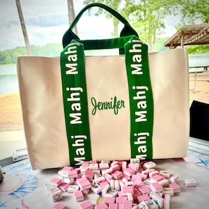 May include: Cream-colored tote bag with green straps and handles. The straps are printed with "Mahj Mahj Mahj" and the name "Jennifer" is embroidered in green. Pink and white Mahjong tiles are scattered in front of the bag.