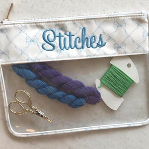 May include: A clear rectangular zippered pouch with a white top border featuring a light blue bow pattern and the embroidered word Stitches in blue. Inside are two small skeins of blue and purple yarn, green embroidery floss, and gold scissors.