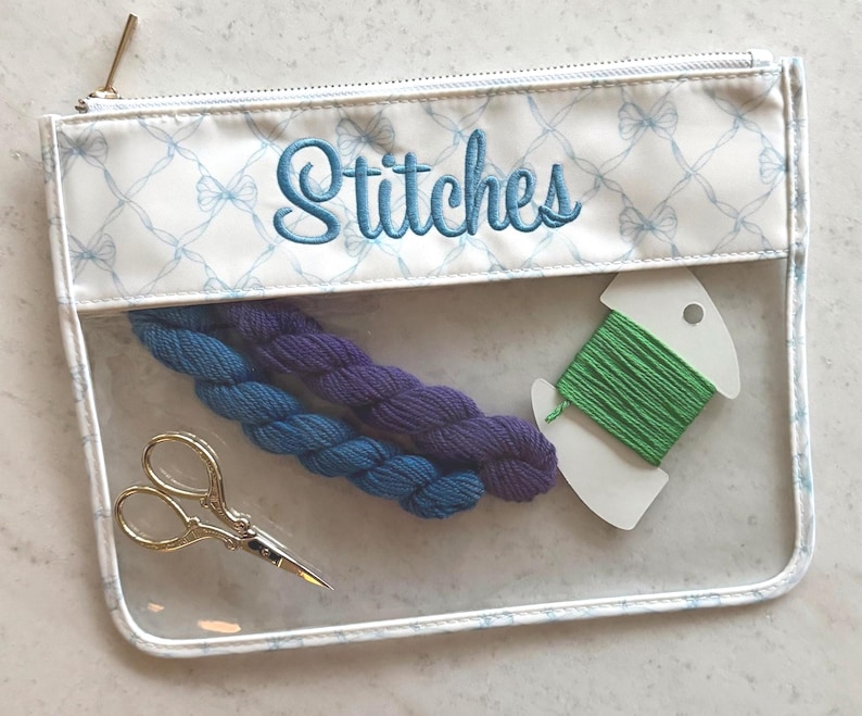 May include: A clear rectangular zippered pouch with a white top border featuring a light blue bow pattern and the embroidered word Stitches in blue. Inside are two small skeins of blue and purple yarn, green embroidery floss, and gold scissors.
