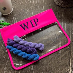 May include: A neon pink rectangular zipper pouch with the text WIP embroidered in dark blue. The pouch features a clear plastic front panel and contains four small skeins of yarn in shades of blue, purple, and lavender inside the bag.