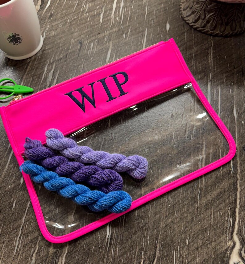 May include: A neon pink rectangular zipper pouch with the text WIP embroidered in dark blue. The pouch features a clear plastic front panel and contains four small skeins of yarn in shades of blue, purple, and lavender inside the bag.