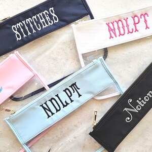 May include: Five rectangular zipper pouches with clear plastic bodies and colored fabric tops. The pouches feature embroidered text including Stitches, NDLPT, and Notions in various colors. Pink and cream embroidery floss are placed near the pouches.