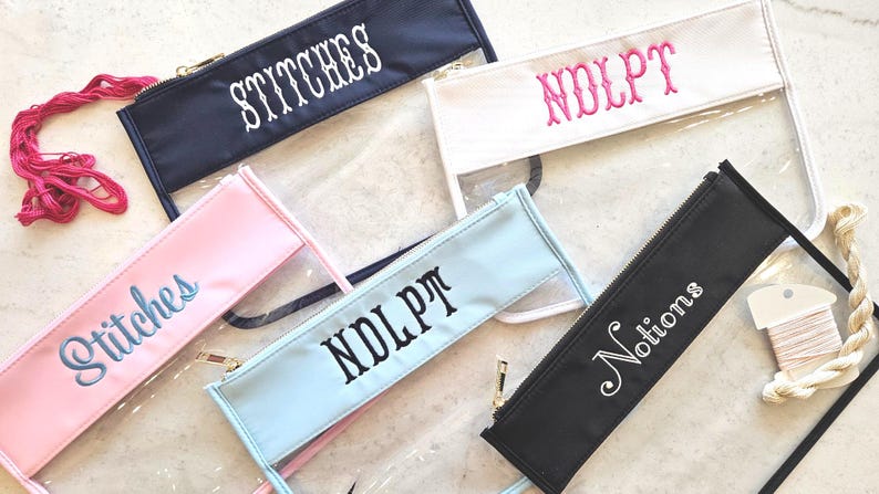 May include: Five rectangular zipper pouches with clear plastic bodies and colored fabric tops. The pouches feature embroidered text including Stitches, NDLPT, and Notions in various colors. Pink and cream embroidery floss are placed near the pouches.