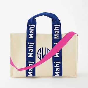 May include: A cream-colored canvas tote bag featuring two blue vertical straps with the repeated white text "Mahj" and a central blue circular monogram logo. The bag includes a bright pink adjustable crossbody strap attached to the sides.