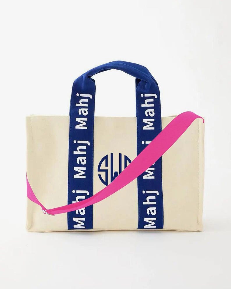 May include: A cream-colored canvas tote bag featuring two blue vertical straps with the repeated white text "Mahj" and a central blue circular monogram logo. The bag includes a bright pink adjustable crossbody strap attached to the sides.