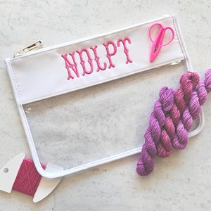 May include: A clear vinyl zipper pouch with white trim and the embroidered pink text "NDLPT". The pouch includes a pair of small pink embroidery scissors and a cluster of variegated purple yarn skeins, plus a pink thread bobbin.