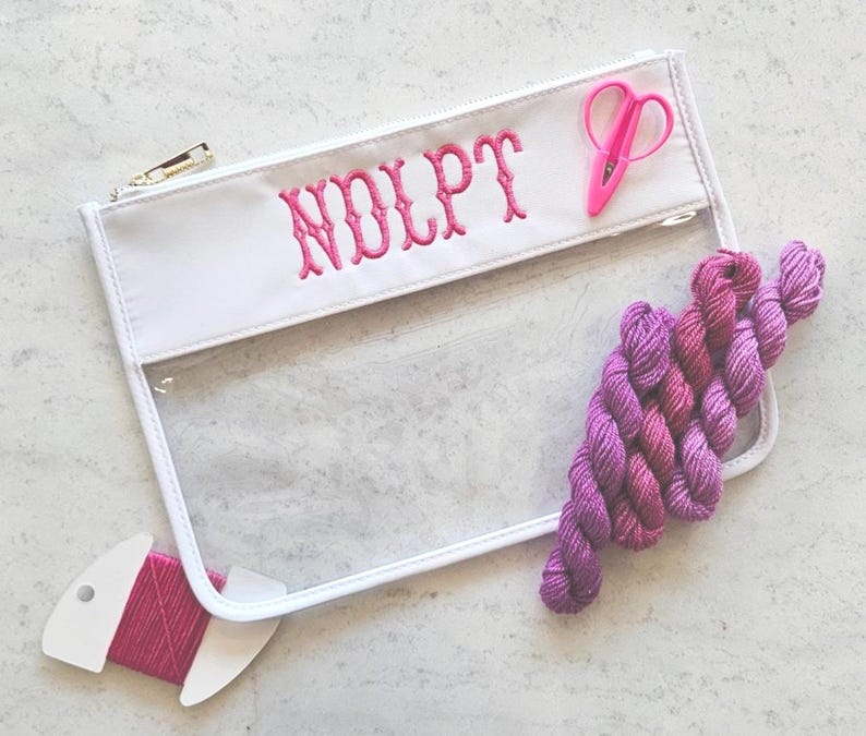 May include: A clear vinyl zipper pouch with white trim and the embroidered pink text "NDLPT". The pouch includes a pair of small pink embroidery scissors and a cluster of variegated purple yarn skeins, plus a pink thread bobbin.