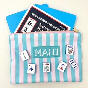May include: A zippered pouch with light blue and pink vertical stripes featuring a rectangular patch that reads MAHJ. The pouch contains a 2025 National Mah Jongg League card and is decorated with six white mahjong tiles and two white dice.