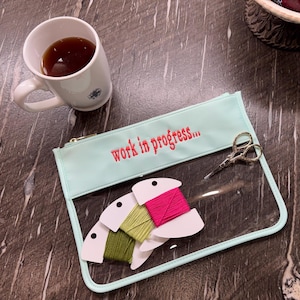 May include: A light mint green zippered pouch with the text "work in progress..." embroidered in red. The pouch has a clear front panel containing three spools of embroidery floss in olive, lime, and hot pink, alongside small silver scissors.