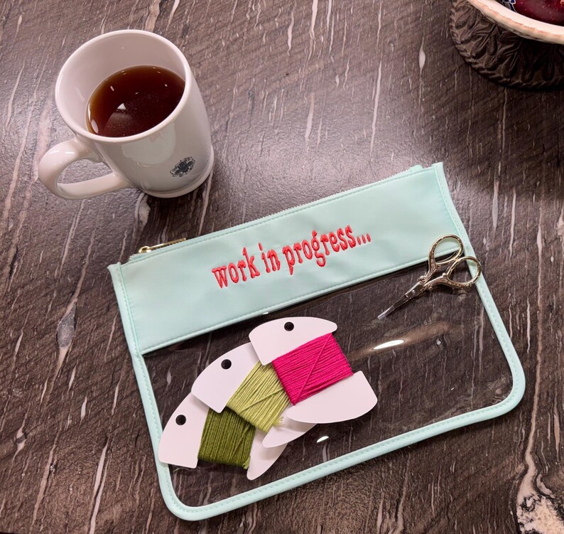 May include: A light mint green zippered pouch with the text "work in progress..." embroidered in red. The pouch has a clear front panel containing three spools of embroidery floss in olive, lime, and hot pink, alongside small silver scissors.