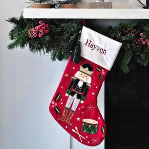 May include: A red Christmas stocking with a white cuff featuring the embroidered name Hayven in dark red. The stocking displays a nutcracker figure, a drum with two drumsticks, and holly leaf accents. A dark green tassel hangs from the top corner.