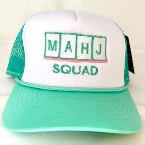 May include: A white and teal trucker hat with the text "MAHJ SQUAD" embroidered on the front. The hat features a white front panel, a teal mesh back, and a teal brim. The letters are in a teal color with a pink outline.