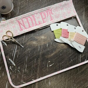 May include: A clear vinyl zipper pouch with a pink and white bow-patterned fabric border. The top fabric panel features the embroidered pink text "NOLPT". Included are gold-toned embroidery scissors and four spools of embroidery floss in pastel colors.