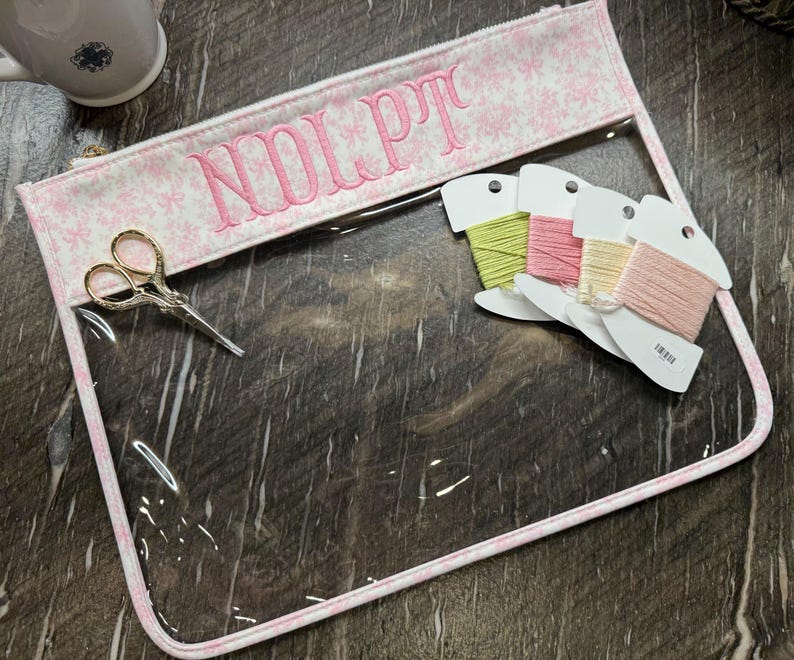 May include: A clear vinyl zipper pouch with a pink and white bow-patterned fabric border. The top fabric panel features the embroidered pink text "NOLPT". Included are gold-toned embroidery scissors and four spools of embroidery floss in pastel colors.