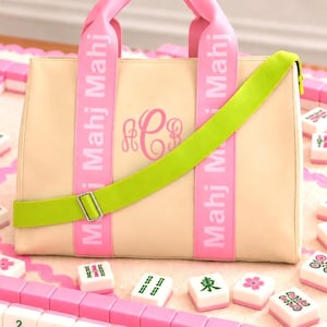 May include: A beige tote bag featuring two pink vertical straps with the white text Mahj Mahj printed on them. The bag has a pink monogram in the center and a bright lime green adjustable crossbody strap with a silver metal buckle attached to the front.