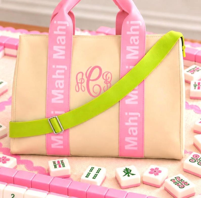 May include: A beige tote bag featuring two pink vertical straps with the white text Mahj Mahj printed on them. The bag has a pink monogram in the center and a bright lime green adjustable crossbody strap with a silver metal buckle attached to the front.
