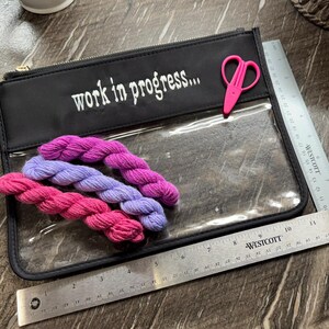 May include: A black zippered pouch with a clear front panel featuring the white text "work in progress..." and a small pink pair of scissors. Three small yarn skeins in shades of pink and purple are inside. A metal ruler shows a width of 11 inches.