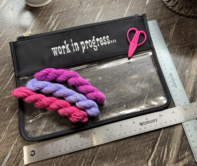 May include: A black zippered pouch with a clear front panel featuring the white text "work in progress..." and a small pink pair of scissors. Three small yarn skeins in shades of pink and purple are inside. A metal ruler shows a width of 11 inches.