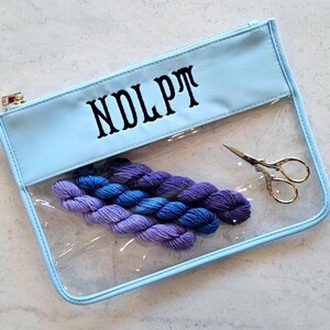 May include: A light blue zippered pouch with a clear front panel and the text NDLPT printed in black. Inside the pouch are four small skeins of purple and blue yarn arranged in a row, alongside a pair of small, ornate gold-toned metal scissors.