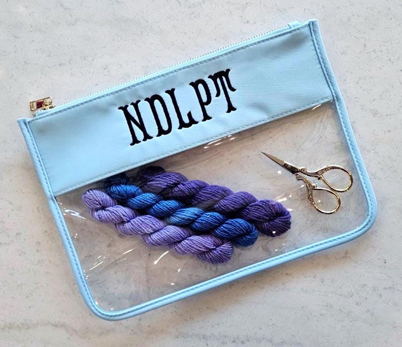 May include: A light blue zippered pouch with a clear front panel and the text NDLPT printed in black. Inside the pouch are four small skeins of purple and blue yarn arranged in a row, alongside a pair of small, ornate gold-toned metal scissors.