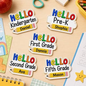 May include: Five embroidered name tags featuring the word HELLO in multi-colored letters, grade levels including Kindergarten, Pre-K, First, Second, and Fifth Grade, and a yellow pencil graphic with personalized names Daniel, Shophia, Dannis, Ava, and Mason.