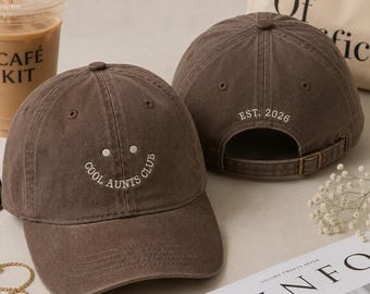Op de afbeelding: Twee donkerbruine baseballpetten van katoen. De voorste pet heeft een geborduurde smiley en de tekst COOL AUNTS CLUB. De achterste pet toont de geborduurde tekst EST. 2026 boven de verstelbare band met een metalen gespsluiting.