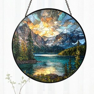May include: Round stained glass window art depicting a mountain lake scene with a sunset. The artwork features a black metal frame and chain for hanging. The landscape includes mountains, trees, and a lake with a colorful sky. The window art is displayed in front of a white curtain.